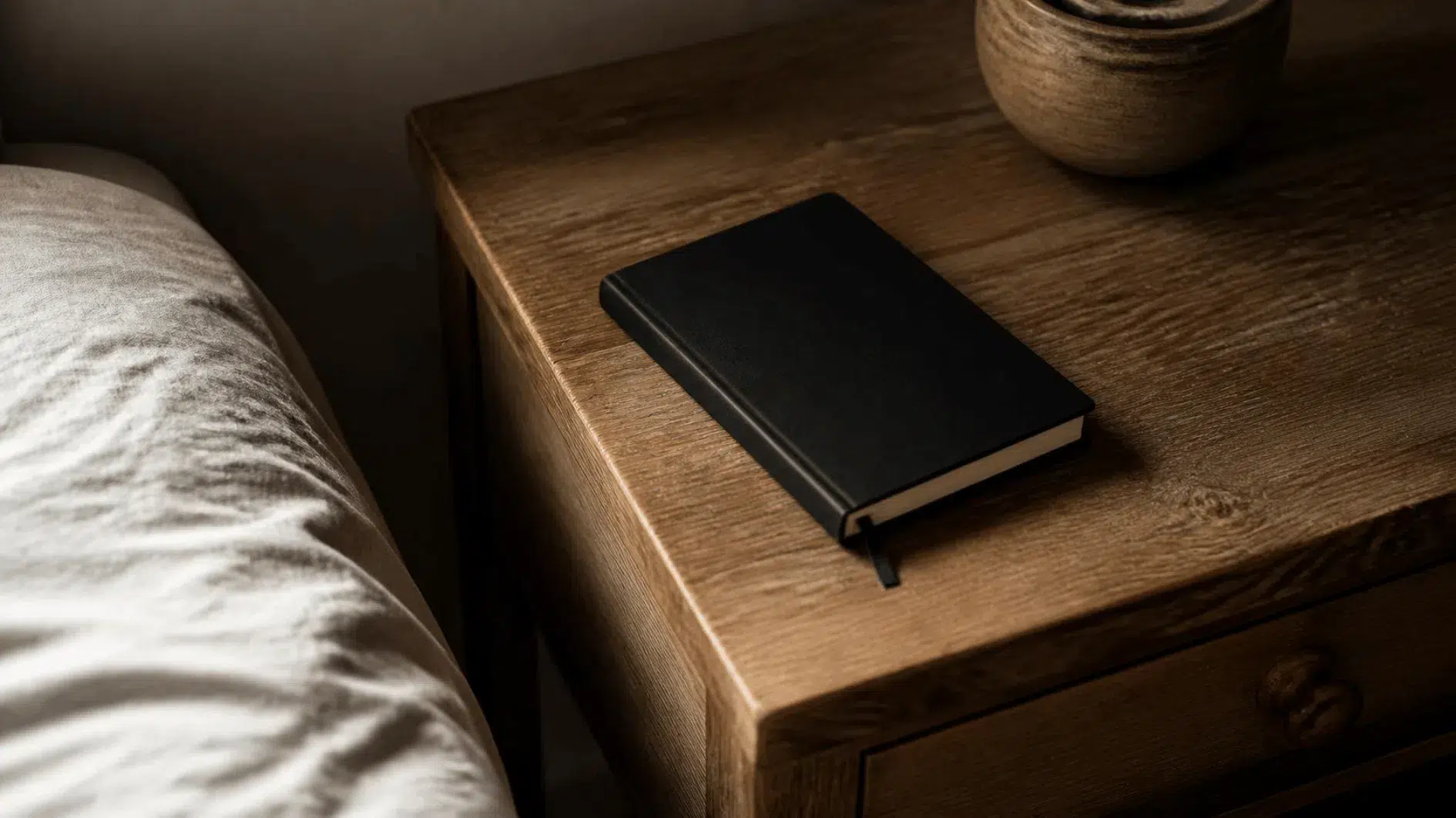 Closed plain linen-bound notebook placed discreetly by a bedside drawer and ceramic lamp